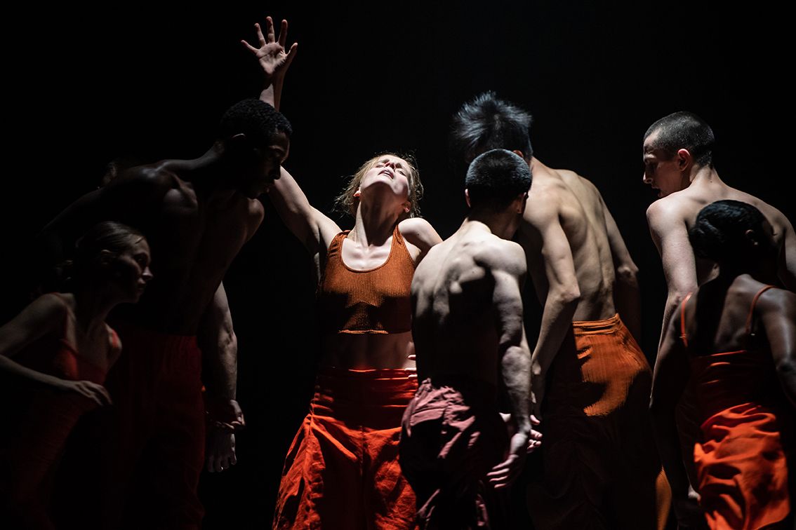 A group of dancers in orange outfits on a dark stage.