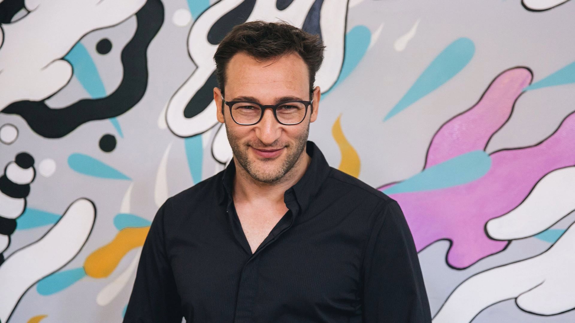 a man wearing glasses standing in front of a colorful wall.