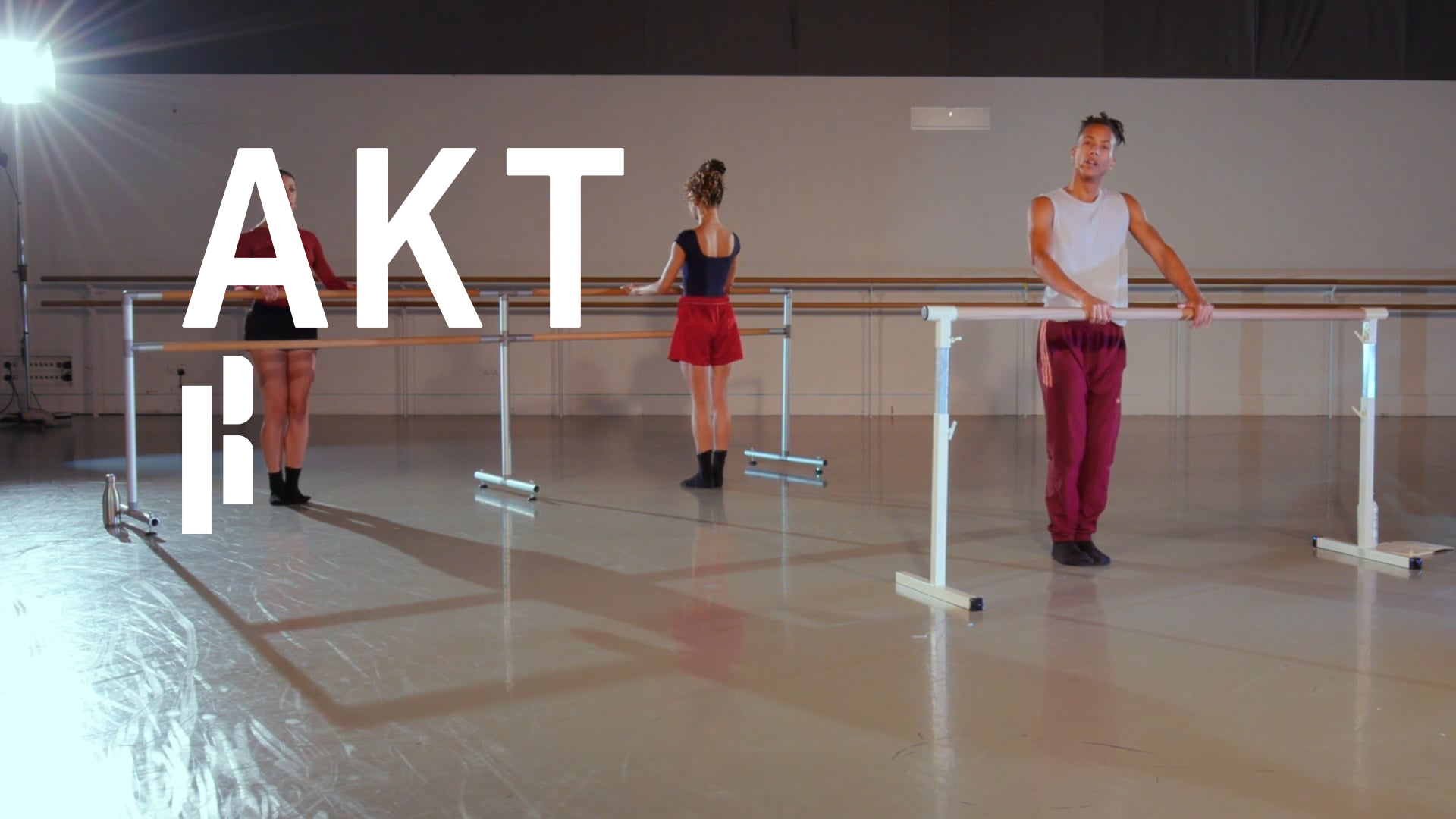 The teacher is holding a barre during an online ballet class. The teacher is a black man wearing red trousers and a grey shirt