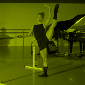 A female ballet dancer with a barre on her left side and a classic piano behind her in-studio