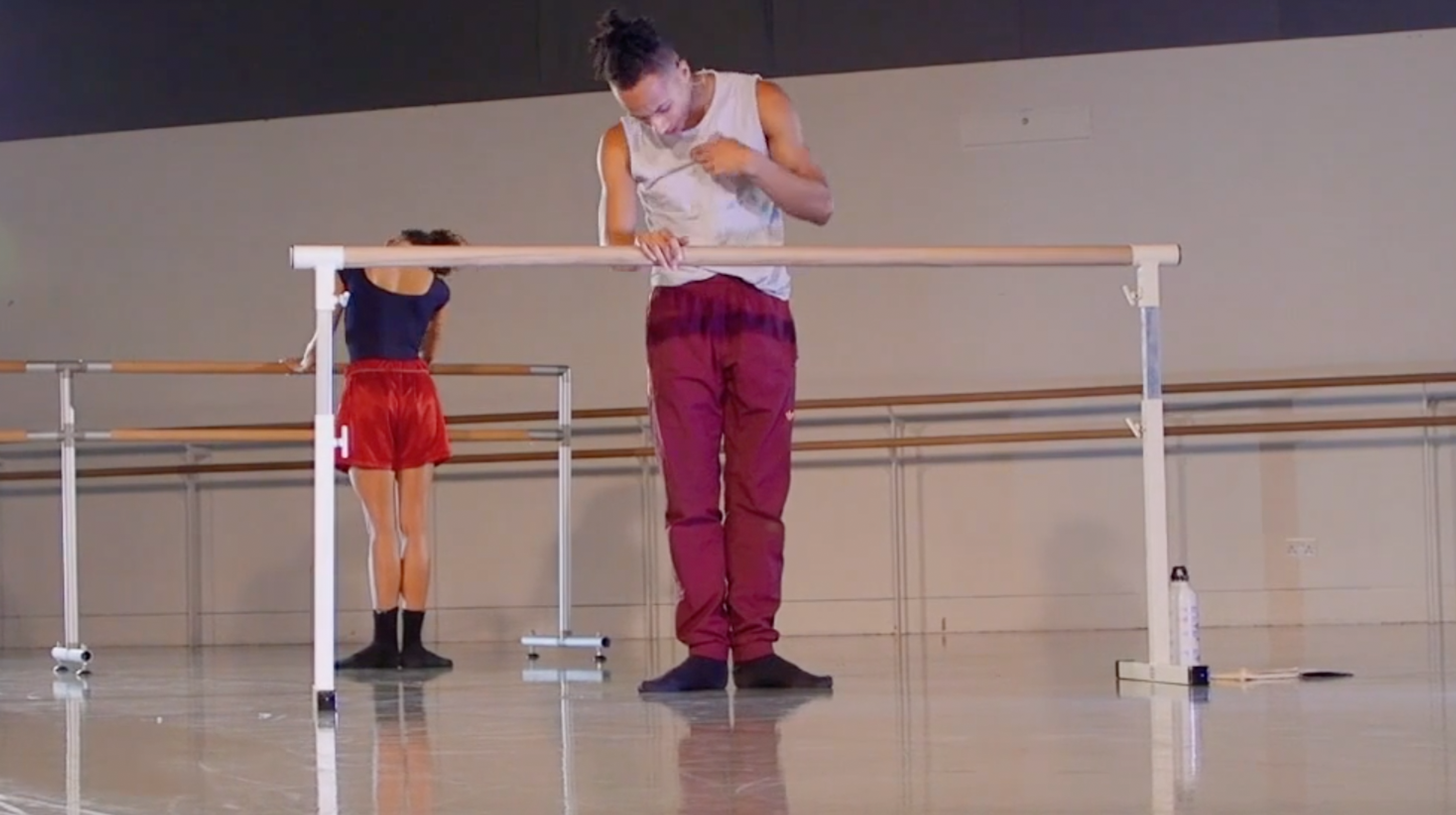 The teacher is holding a barre during an online ballet class. The teacher is a black man wearing red trousers and a grey shirt