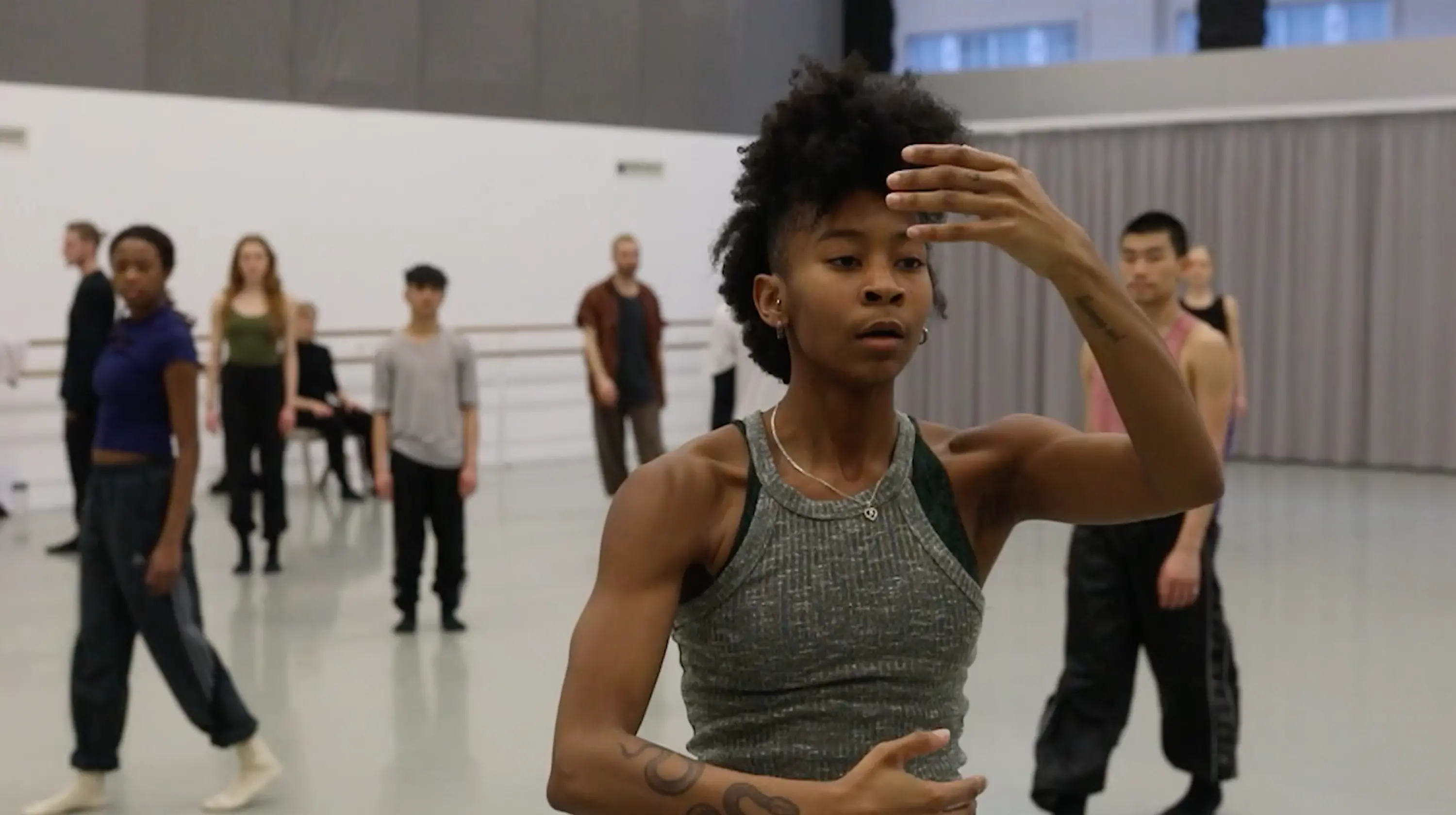 Rambert dancer, a black woman with curly hair, at the centre of the studio with a group of other dancers behind her.