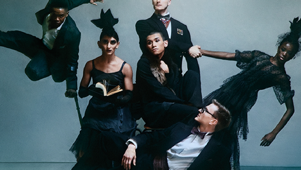 A group of six individuals in formal and avant-garde outfits are posed in a dynamic fashion against a grey background; some are standing, sitting, or mid-air.