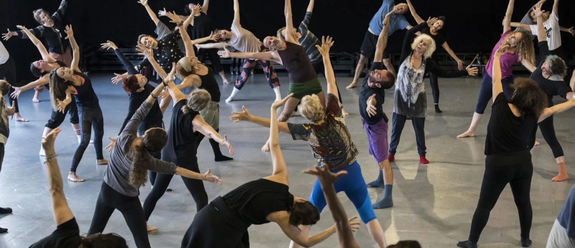 A group of people are engaged in a dance class, performing various expressive movement poses on a gray studio floor. They are barefoot and dressed in casual dance attire.