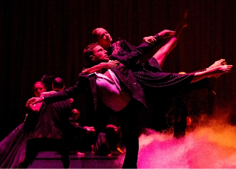 Two dancers perform a contemporary dance, one lifting the other in a dramatic pose against a dark, smoky background with pink and purple lighting. Other performers are visible in the background.