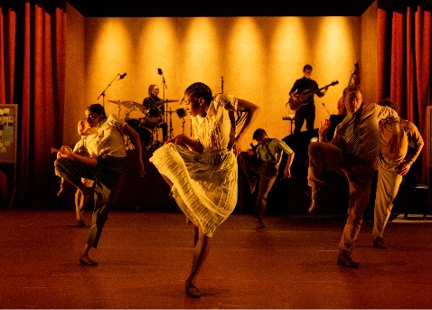 A group of dancers perform energetically on stage with a band playing live music in the background. The stage is lit with warm, dim lighting.