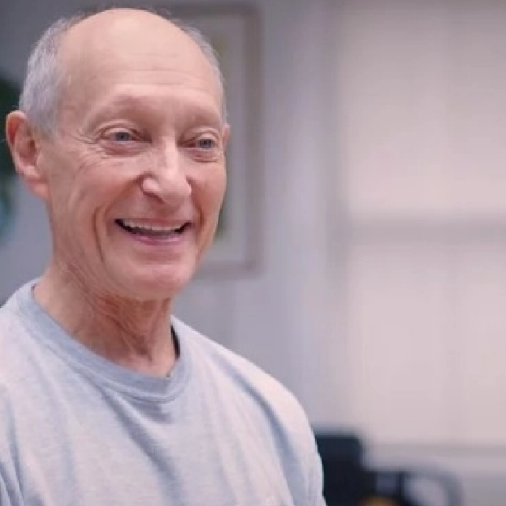 An older man wearing a gray shirt smiles while standing indoors near a large plant.