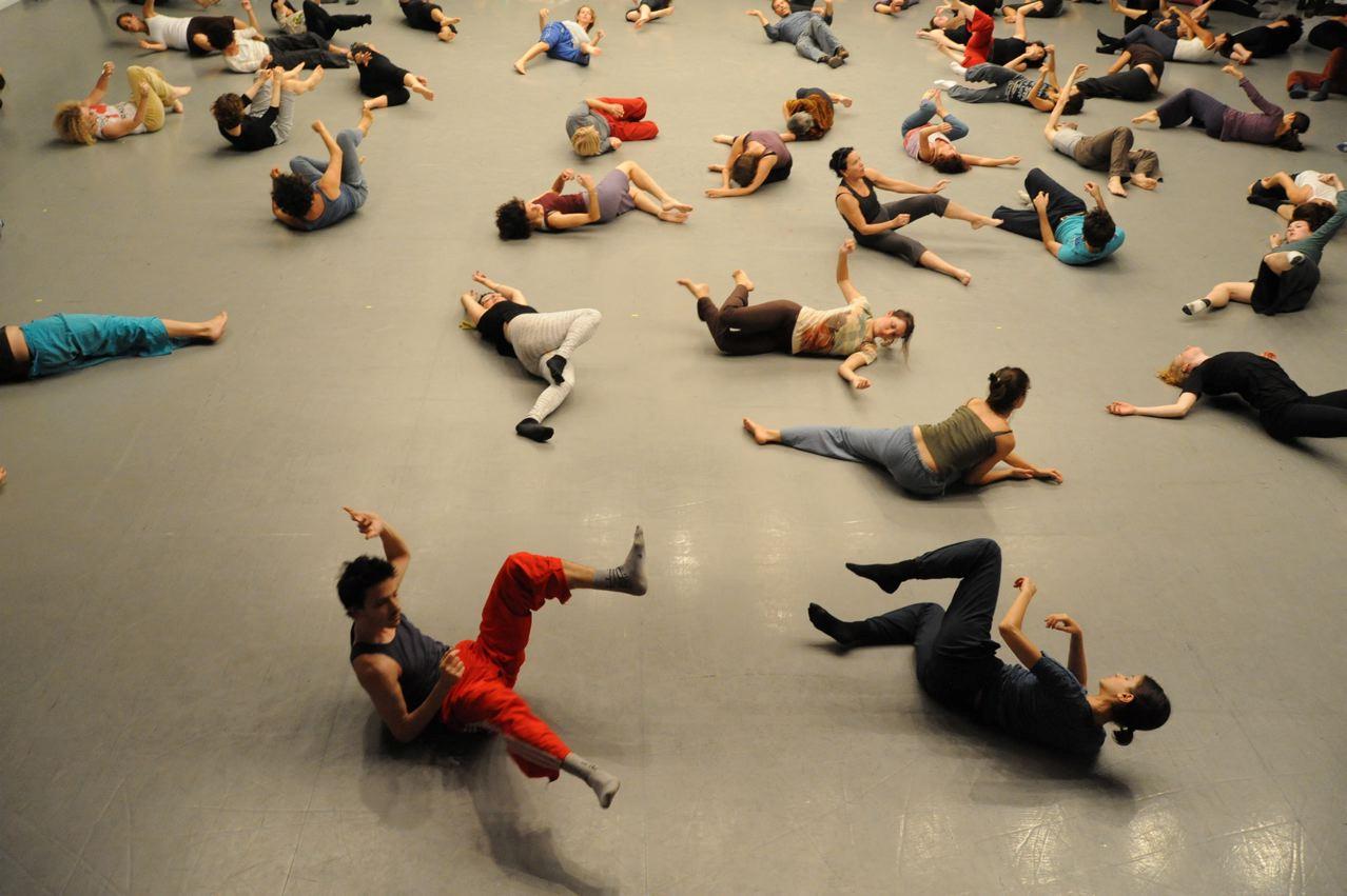A group of people lying on a gray floor, engaged in various movements and poses, suggesting a dance or movement class.