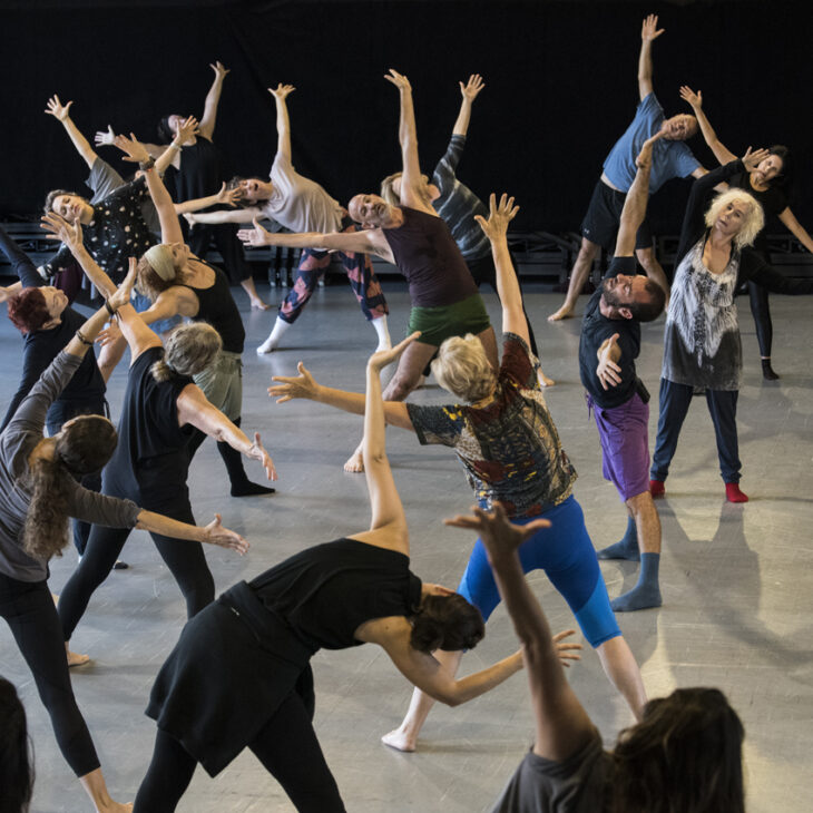 A group of people in a dance studio engage in a stretching exercise, extending their arms and tilting their heads back.