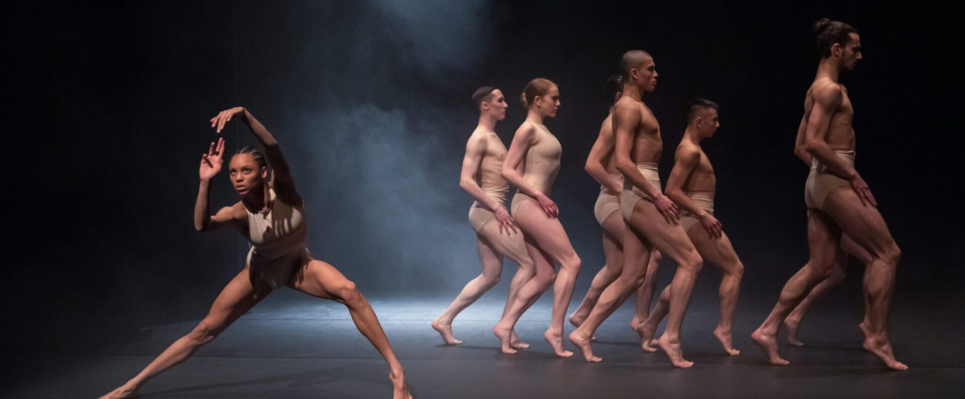 A group of six dancers in minimal costumes perform a contemporary dance on a dimly lit stage, with one dancer extending her arms and legs in the foreground and the others walking in line behind her.