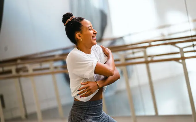 A person wearing a white top and gray leggings is smiling and stretching arms in a brightly lit room with mirrored walls and ballet barres.