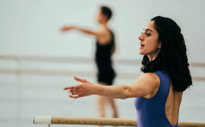 Woman in a purple leotard instructing in a dance studio with another person in the background.