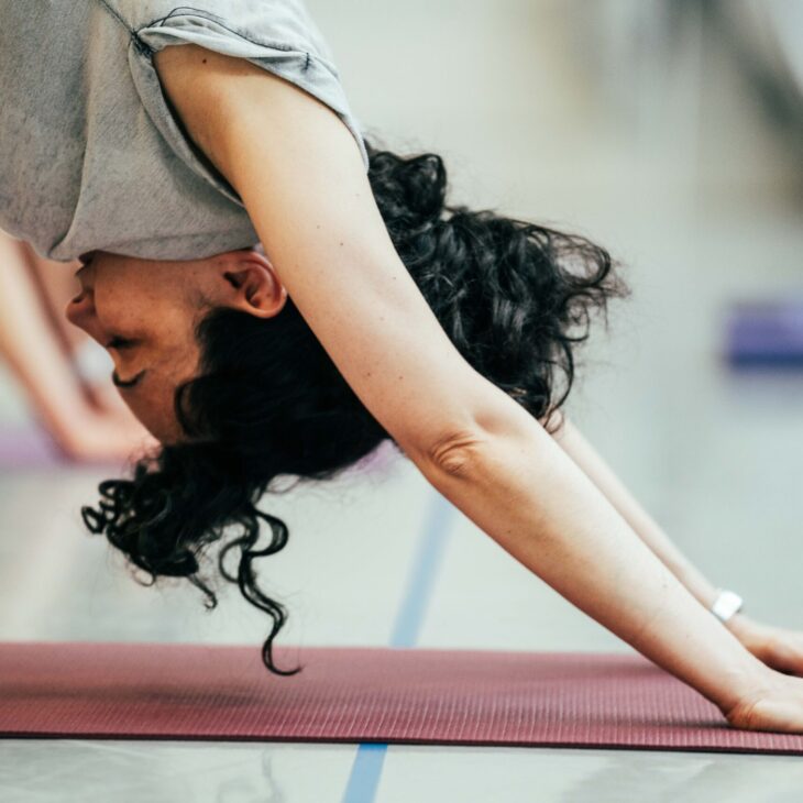 Person practicing yoga, performing a downward-facing dog pose on a purple mat in a room with others in the background.