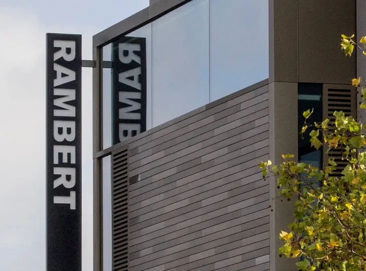 Modern building with reflective glass and "RAMBERT" signage. Gray and wooden exterior with a tree on the right side. Sky in the background.