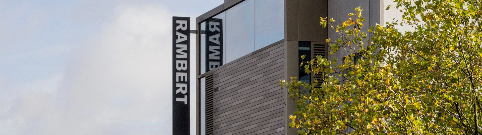 Modern building with reflective glass and "RAMBERT" signage. Gray and wooden exterior with a tree on the right side. Sky in the background.