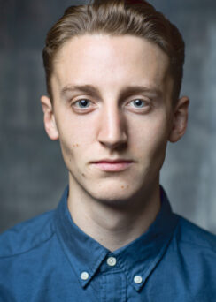 A young man with short, light brown hair and blue eyes wearing a blue button-up shirt looks directly into the camera against a neutral background.