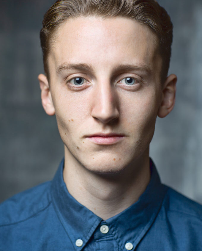 A young man with short, light brown hair and blue eyes wearing a blue button-up shirt looks directly into the camera against a neutral background.
