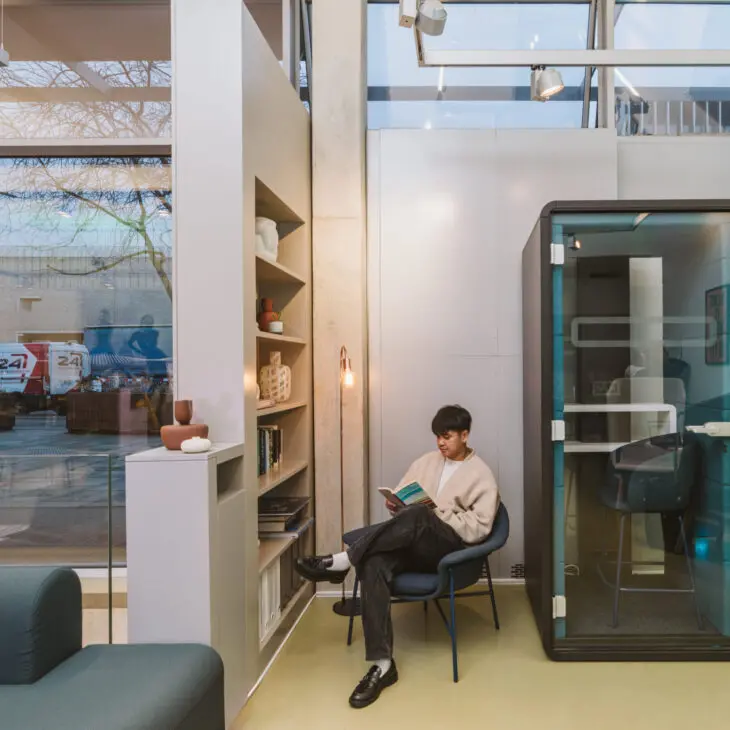 A person sits in a modern office space reading a book next to a bookshelf and a small enclosed booth. The setting is illuminated by natural light from large windows.