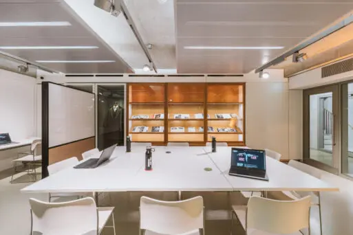 A modern office meeting room with white tables, chairs, open laptops, water bottles, and bookshelves displaying documents in the background. Ceiling lights illuminate the space.
