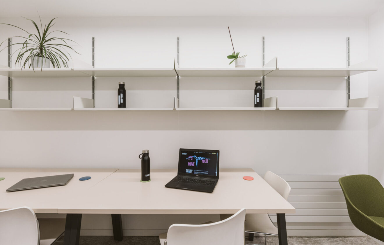 A minimalist office setup with two desks, a laptop, two water bottles, and a potted plant. The background features white shelves with a few items, including another plant and additional water bottles.
