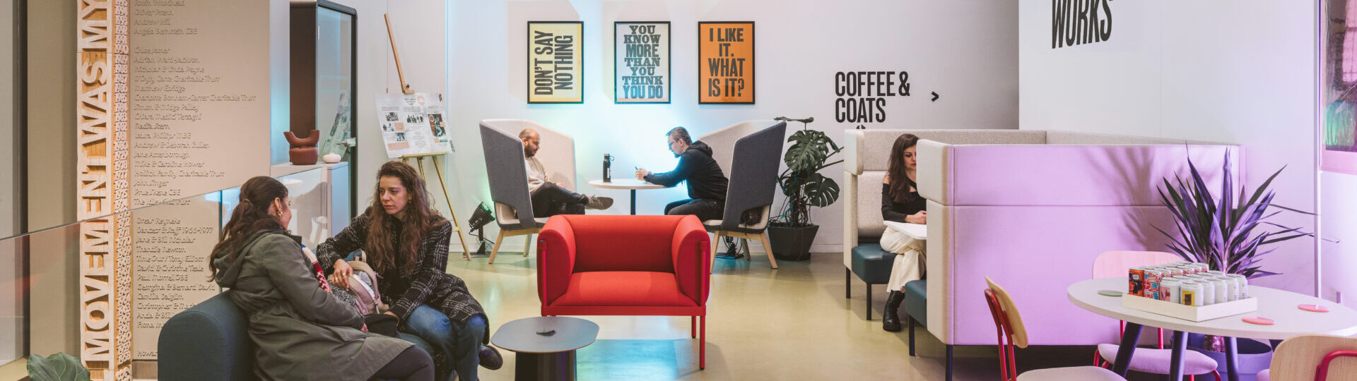 A modern cafe with people sitting and conversing at various tables and booths. Signs read "Rambert Works," "Coffee & Coats," and others displaying motivational phrases.