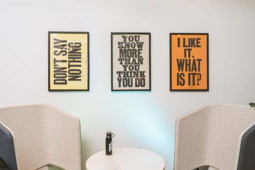 Three framed motivational posters on a wall above a white round table with a black water bottle. Two light gray chairs are positioned on either side of the table.