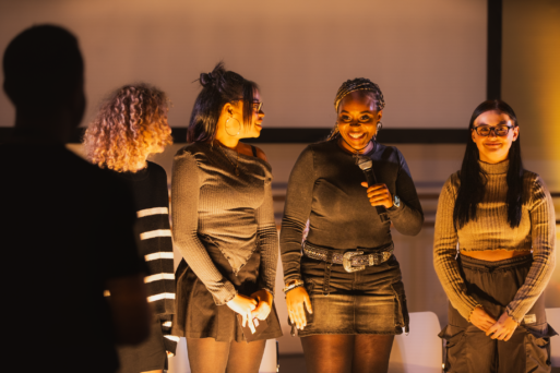 A group of four women standing on a stage, with one woman holding a microphone and speaking. They all appear to be engaged in the event.