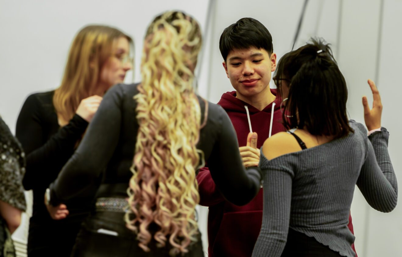 A group of young adults is engaged in conversation. One person with long curly hair is in the foreground, while others are gathered around and interacting.
