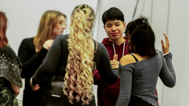 A group of young adults is engaged in conversation. One person with long curly hair is in the foreground, while others are gathered around and interacting.
