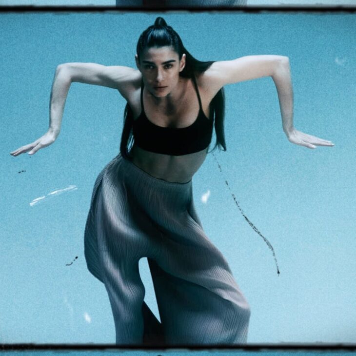 A person with long hair in a ponytail and wearing a black top and loose pants is in mid-dance, arms bent outwards, against a blue background.