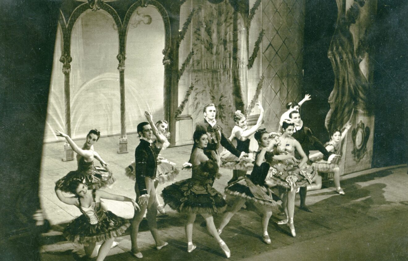 A group of ballet dancers performs on stage in intricate costumes, with a detailed backdrop and theatrical lighting.