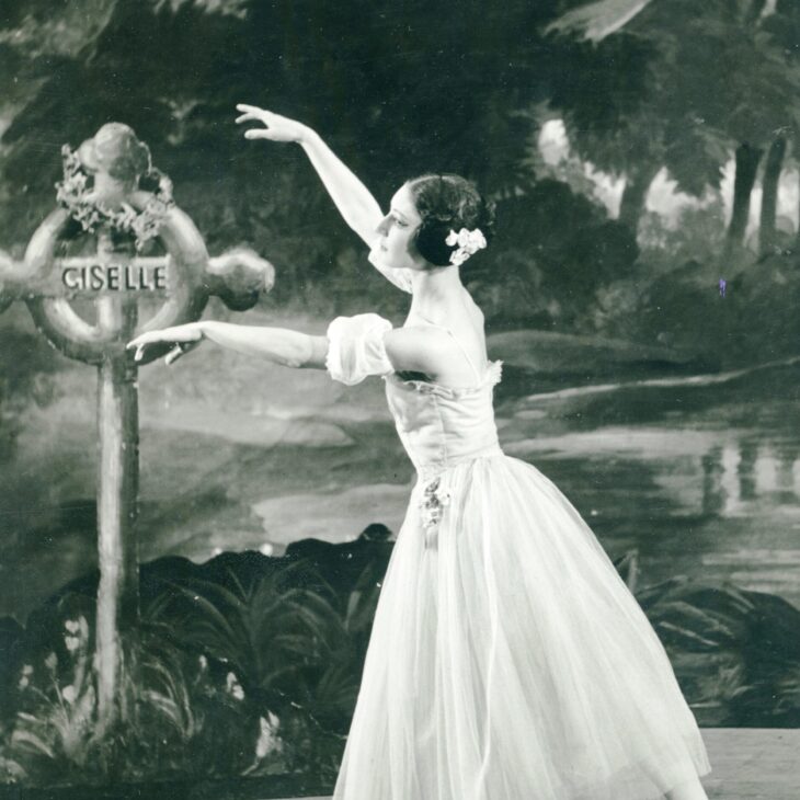 A ballerina in a tutu performs on stage, balancing on one foot in front of a sign that reads "Giselle" with a woodland backdrop.