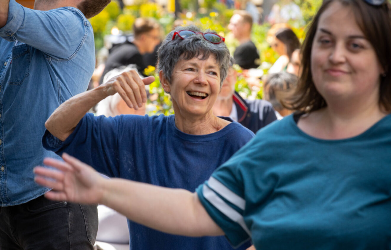 A group of people outdoors, smiling and dancing together in casual clothing. The background shows trees and a small crowd.