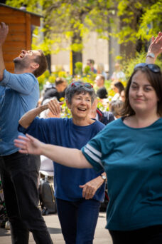 A group of people outdoors, smiling and dancing together in casual clothing. The background shows trees and a small crowd.