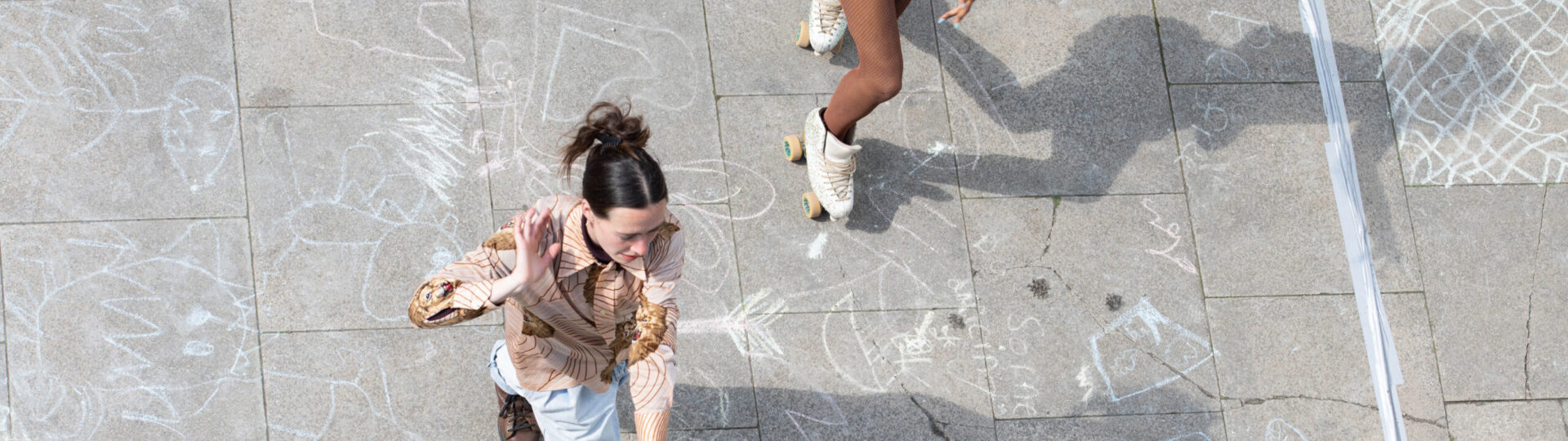 Two people dressed in bright outfits roller skate on a chalk-drawn pavement, viewed from above. The ground is decorated with various chalk drawings.