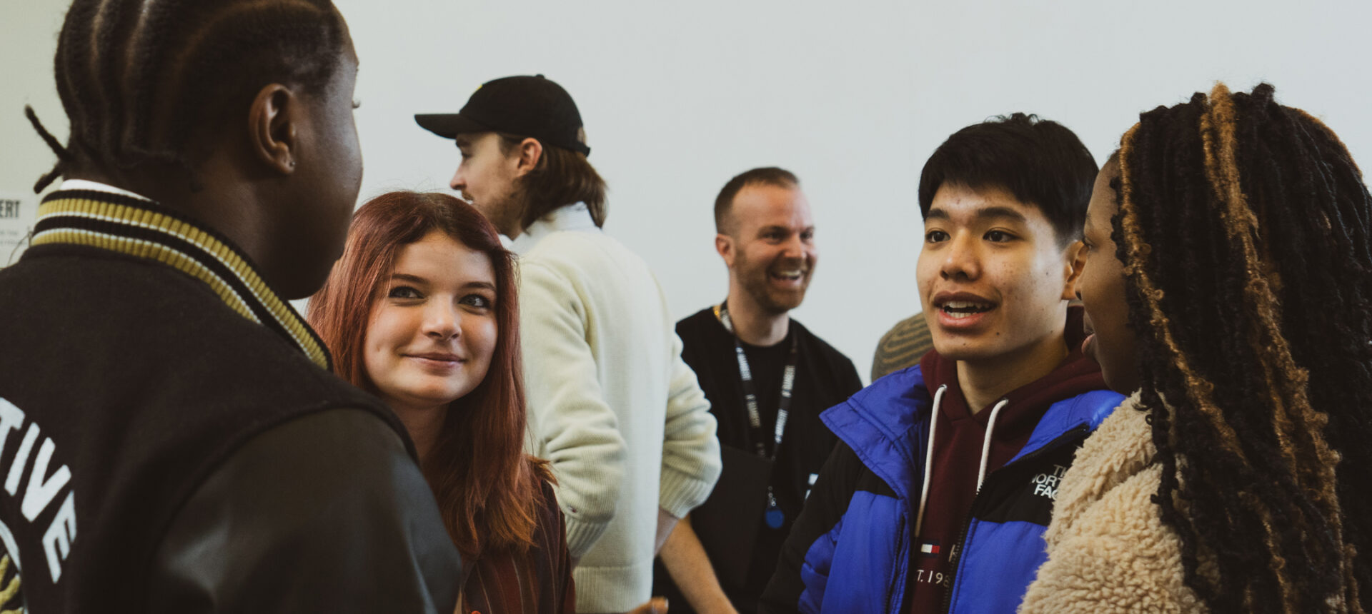 A group of people engaged in conversation indoors; one person wears a black and white jacket, another with red hair smiles, and a third person in a blue jacket speaks to another wearing a tan coat.