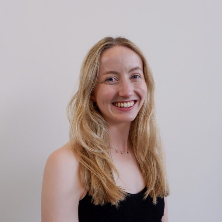 A woman with long blonde hair and a black top smiles at the camera against a plain white background.