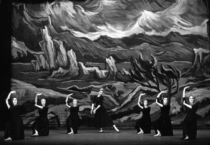 A group of dancers in dark, flowing garments perform against a dramatic backdrop depicting a rugged landscape with mountains and twisted trees.