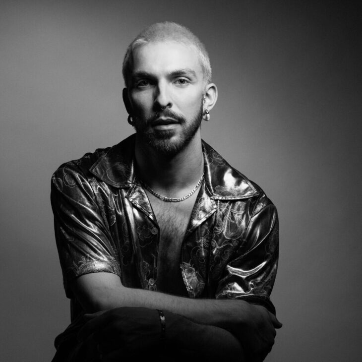 A black and white portrait of a person with short light hair and dark facial hair, wearing a dark button up shirt, set against a plain background.