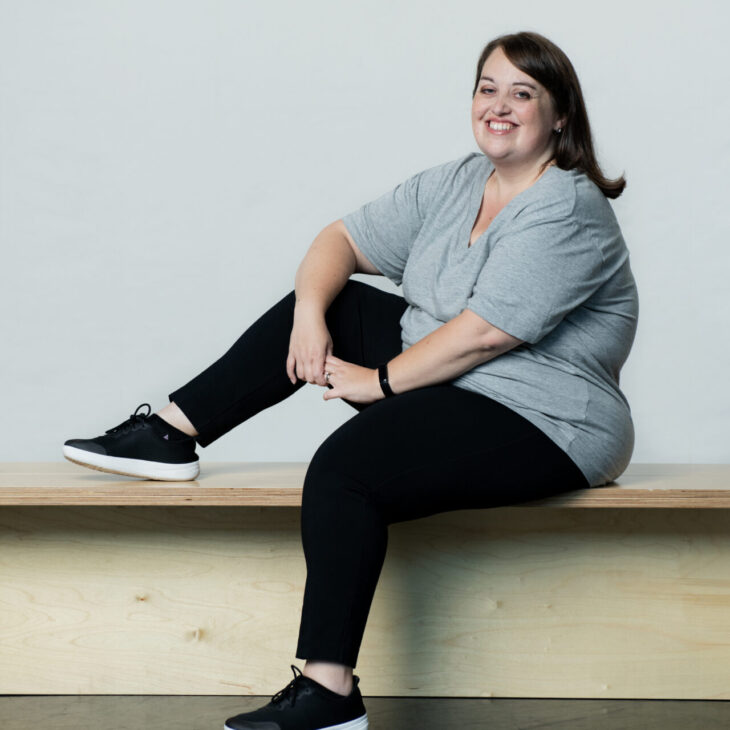 A person in a gray shirt and black pants, wearing black sneakers, sits on a wooden bench with one leg up and smiles at the camera.