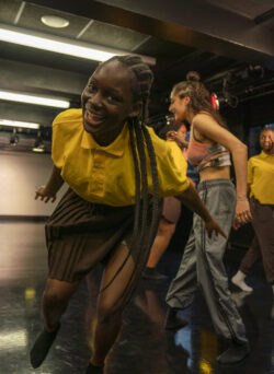 A person wearing a yellow shirt and brown skirt smiles and leans forward while dancing in a studio, with others dancing in the background.