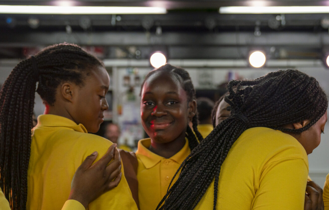 Four people wearing yellow shirts, with braided hairstyles, standing closely together indoors. One person in the center is looking at the camera and smiling while the others are turned away or looking down.