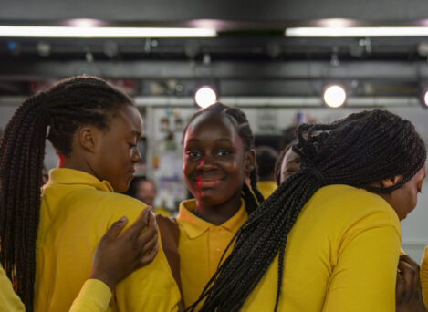 Four people wearing yellow shirts, with braided hairstyles, standing closely together indoors. One person in the center is looking at the camera and smiling while the others are turned away or looking down.