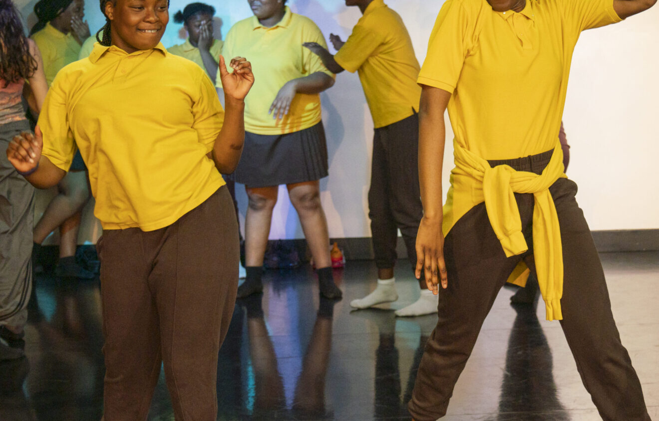 A group of people in yellow shirts and dark pants dance energetically on a stage with a polished floor.