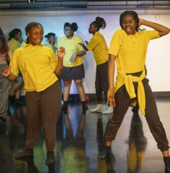 A group of people in yellow shirts and dark pants dance energetically on a stage with a polished floor.