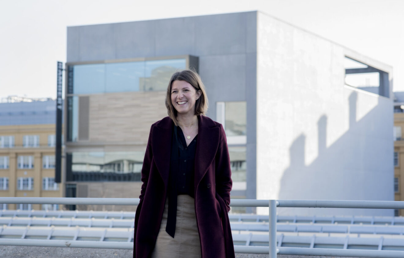 A woman with shoulder-length hair in a maroon coat and black shirt stands in front of a modern building, smiling.