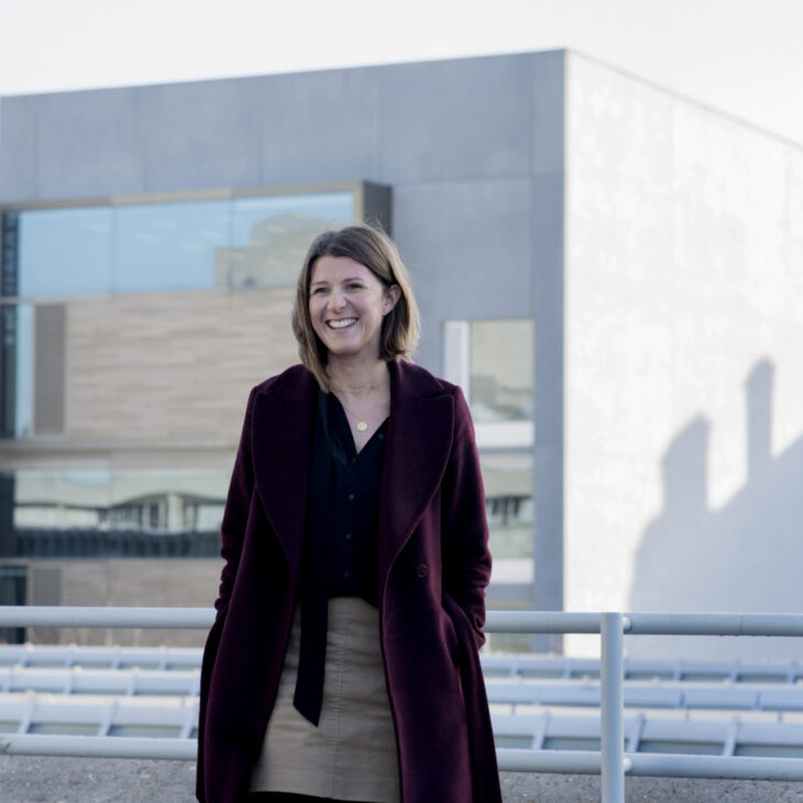 A woman with shoulder-length hair in a maroon coat and black shirt stands in front of a modern building, smiling.