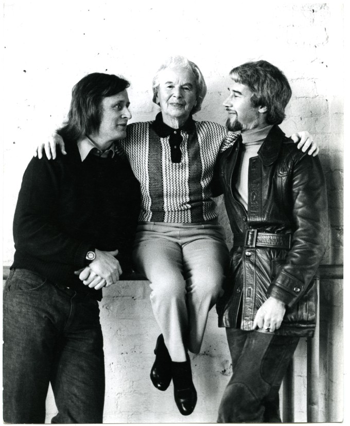 Three people pose for a photo against a white brick wall. The seated person in the center is flanked by two people standing on either side, one wearing a dark sweater and the other in a leather jacket.