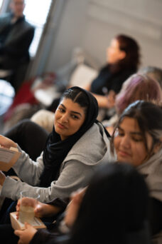 Several women sitting together in a room, engaged in conversation. One woman is holding a book and another a glass.