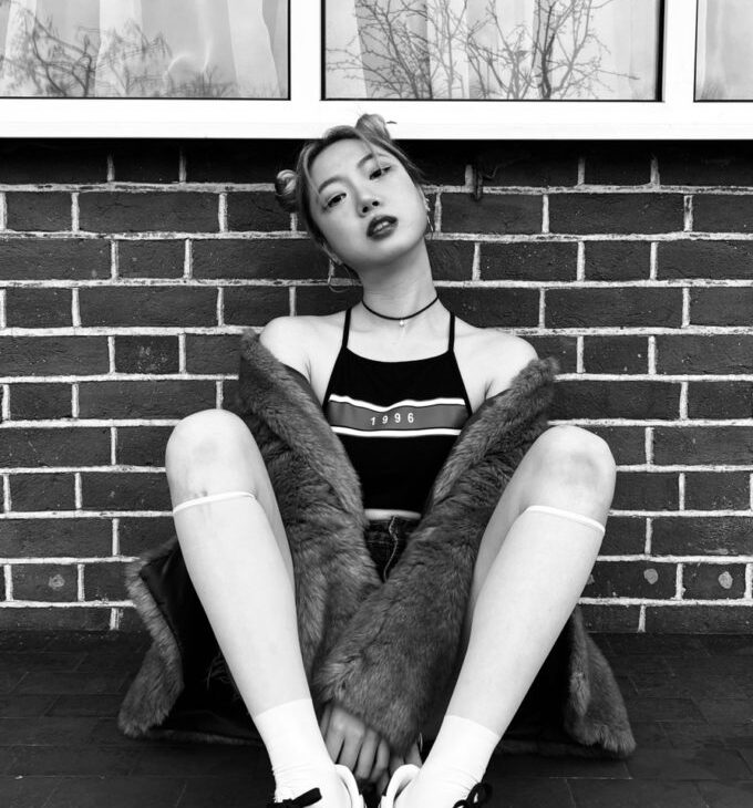 A person wearing a striped top, fur coat, and sneakers sits against a brick wall with a window behind them. They have a serious expression, and their hair is styled in buns. The photo is in black and white.